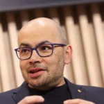 Sir Demis Hassabis em entrevista, expressão séria, fundo desfocado