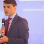 Hugo Valadares, diretor do MCTI, discursando em evento oficial com bandeira do Brasil ao fundo.