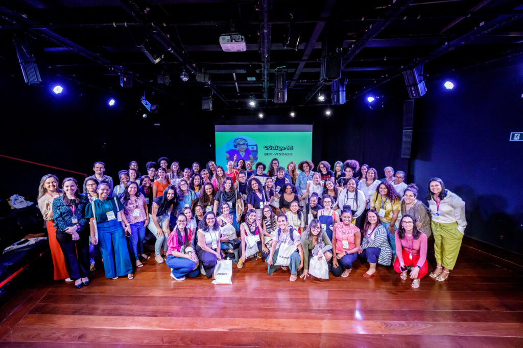 Participantes de diferentes idades e etnias em oficina do projeto Código M com banner da Laboratória ao fundo