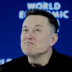 Elon Musk em conferência com telas mostrando logos da Tesla e X/Grok ao fundo