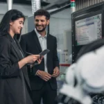 Profissional trabalhando em laptop com gráficos e sensores IoT em ambiente industrial ao fundo