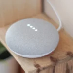 Google Home Mini e smartphone mostrando onda sonora do assistente, com papéis judiciais ao fundo